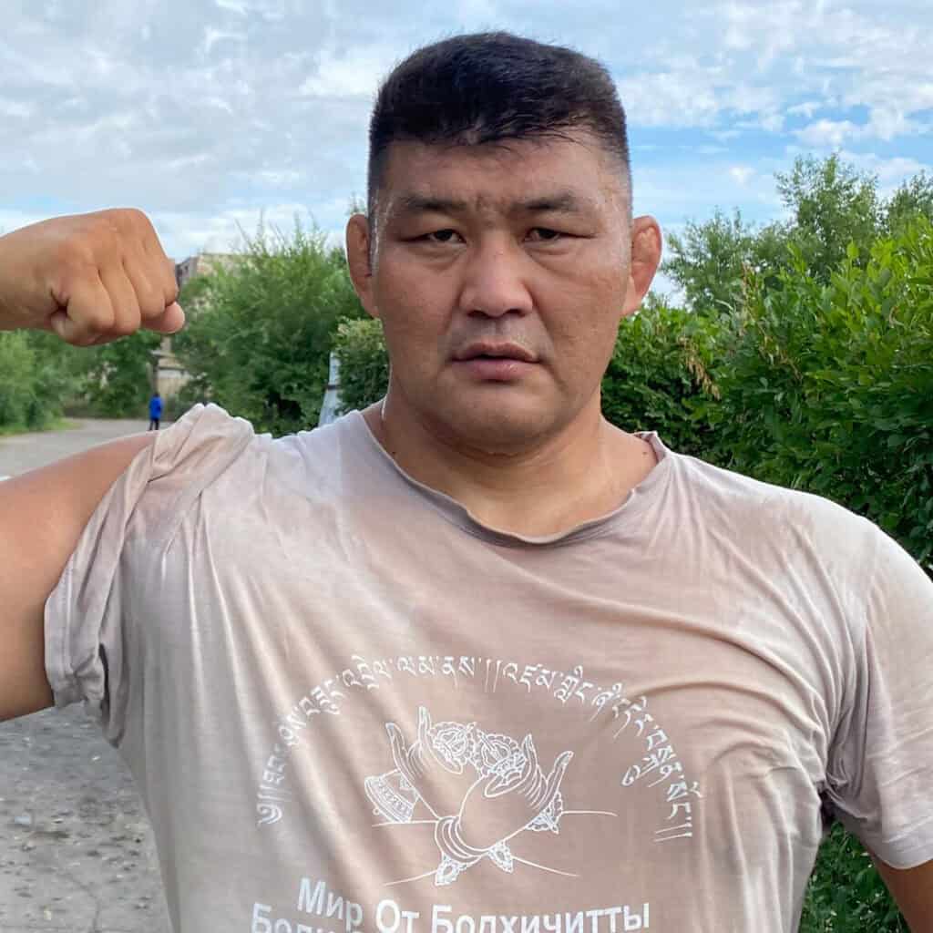 Sumo wrestler showing muscle outdoors with greenery and cloudy sky.