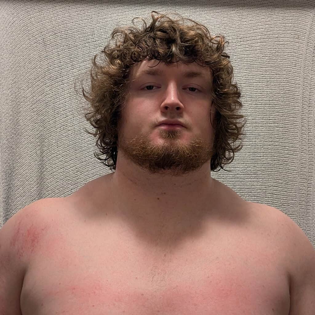 Young man with curly hair and beard, shirtless, against a textured background.