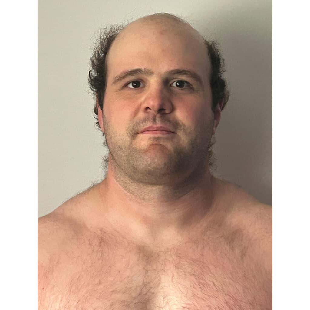 Man with short hair and a serious expression, shirtless, against a plain background.