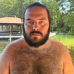 Man with dark hair and beard looking at camera outdoors.
